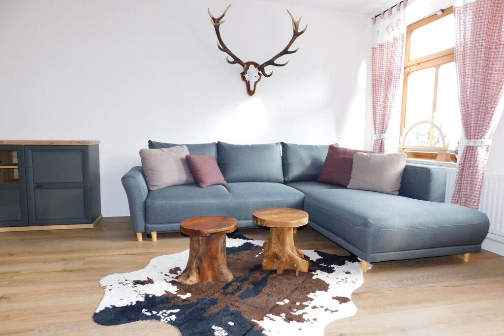 Un salon moderne avec un canapé d’angle, deux tabourets en bois, un tapis en peau de vache et une tête de cerf fixée au mur.