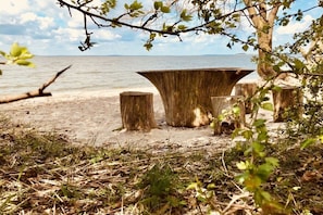 Ein Strandambiente mit einem großen Holztisch und zwei Hockern, umgeben von Bäumen und Vegetation.