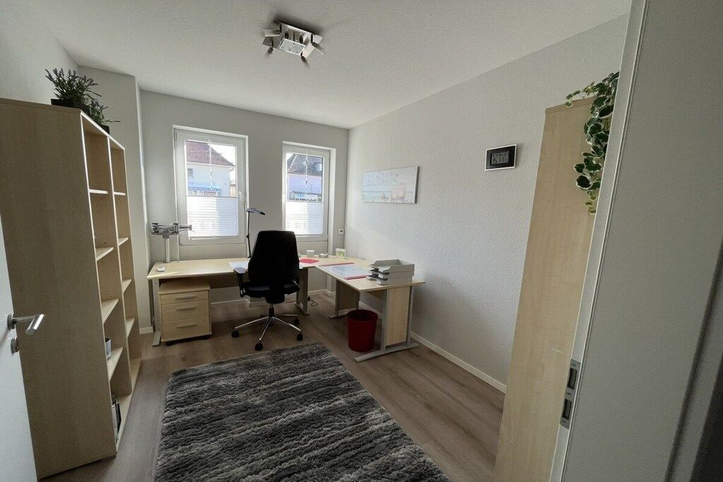 Una habitación con escritorio, silla y estantería. Hay una ventana con persianas, una puerta y una planta pequeña en la pared.