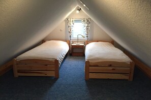 Een compacte slaapkamer op zolder met twee houten bedden, een raam met gordijnen en een bureau met lamp.
