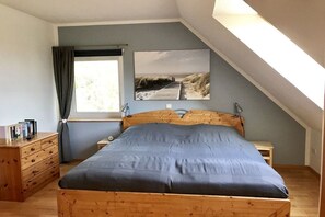 A bedroom with a wooden bed, a nightstand, a dresser, a window with curtains, and a beach-themed wall art. - Holiday apartment with 3 bedrooms (Wangerland)