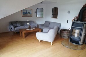 Een woonkamer met een bank, fauteuil en een klein tafeltje. Er is een open haard met schoorsteen en een raam met uitzicht op een gebouw.