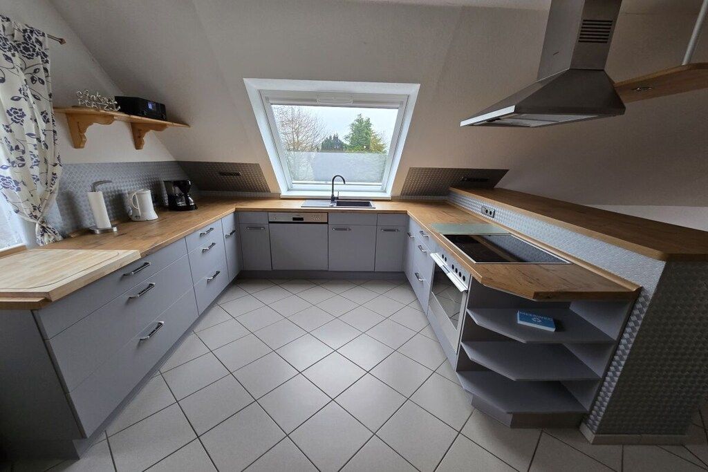 Eine kleine Küche mit grauen Schränken, einer Holz Arbeitsplatte, einem eingebauten Backofen und einem Fenster über der Spüle.