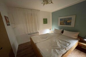 Un dormitori amb un llit de fusta, roba de llit blanca, un radiador, una finestra amb cortines i un quadre penjat a la paret.