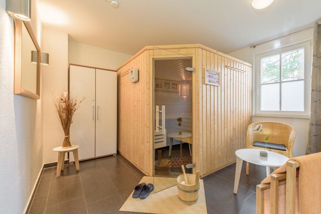 Una sauna con separador de madera, una mesita con una planta, un par de pantuflas y un cubo con una cuchara.