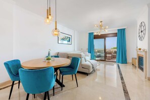 A modern living room with a round dining table, blue chairs, a sofa, and a large window with curtains. - Finca San Antonio Mijas II – Sea View Apartment with Pool & Terrace in Mijas Pueblo (Mijas Pueblo)