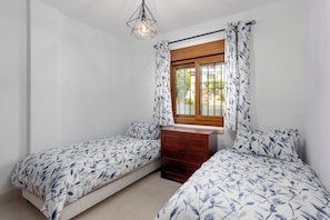 A bedroom with two beds, a wooden dresser, a window with curtains, and a hanging light fixture. - Finca San Antonio Mijas II – Sea View Apartment with Pool & Terrace in Mijas Pueblo (Mijas Pueblo)