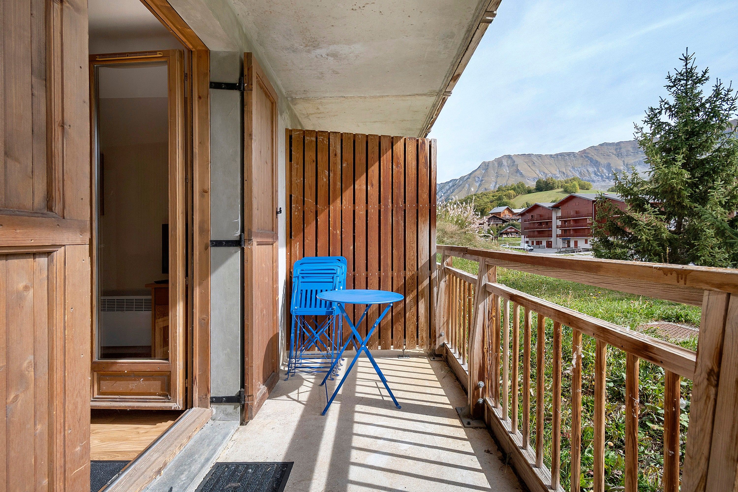A wooden balcony with a blue folding chair and a small table, overlooking a mountainous landscape with buildings.