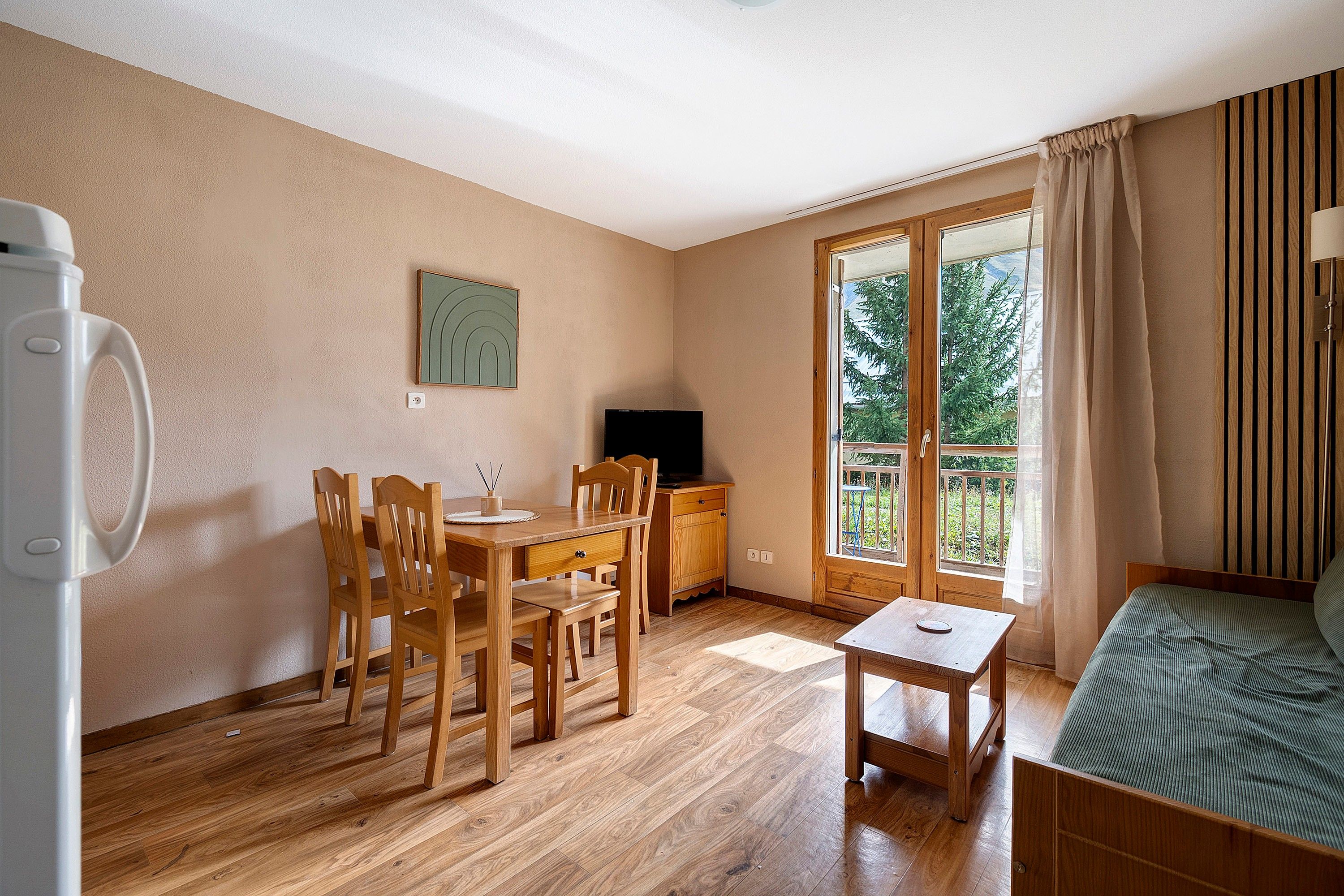 Een kamer met een houten eettafel, stoelen, een klein bijzettafeltje, een televisie, een bank en een groot raam met gordijnen.
