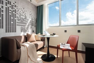 A modern hotel room with a patterned wall, a sofa, a small table with a teapot, a chair, and a window with a view of the sky.