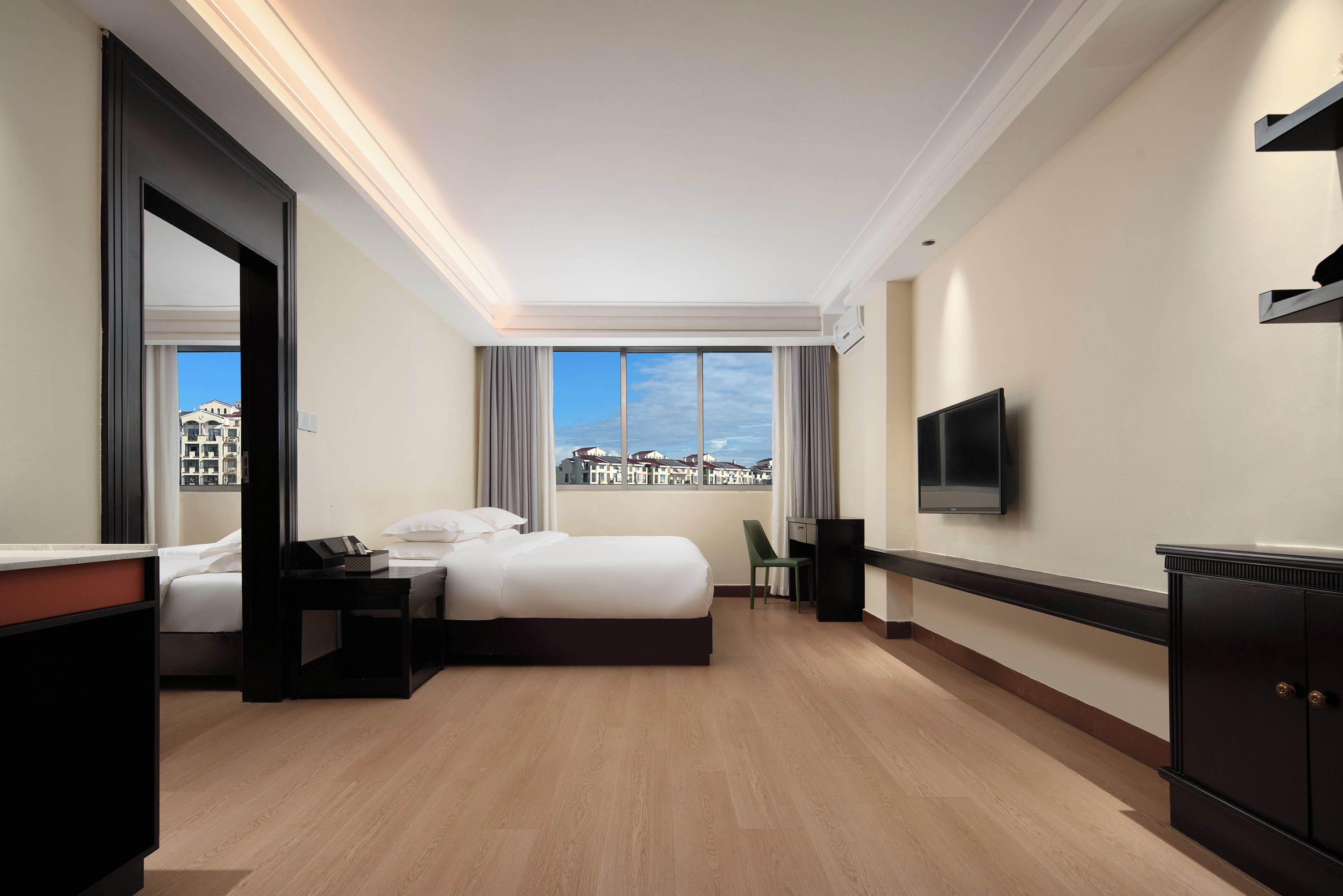A hotel room with a bed, bedside table, chair, TV, and a view of buildings through the window.