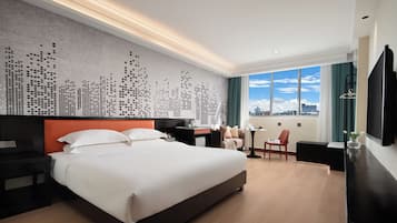 A hotel room with a large bed, a desk, a chair, a TV, and a cityscape view through the window.
