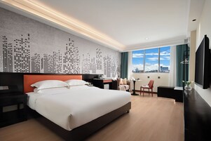 A hotel room with a large bed, a desk, a chair, a TV, and a cityscape view through the window.