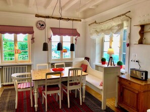 Un coin repas chaleureux avec une table en bois, des chaises blanches et un bol rouge. On y trouve des fenêtres avec des rideaux à carreaux, un radiateur et une petite étagère avec des bocaux.