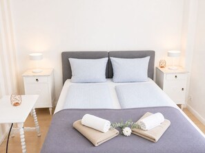 Un lit double bien fait, avec une tête de lit grise et des oreillers assortis, deux tables de chevet avec des lampes, et une petite table sur laquelle est posé un objet décoratif.