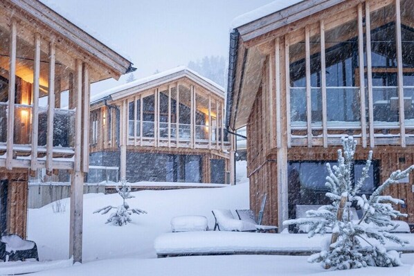 A wooden cabin with large windows, a covered outdoor area, and a snow-covered landscape.