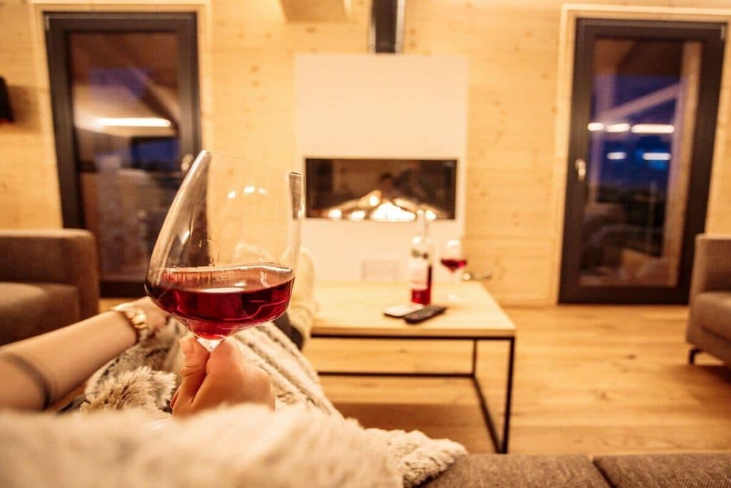 Eine Person hält ein Glas Rotwein in einem gemütlichen Wohnzimmer mit Kamin, Couchtisch und Sofa.