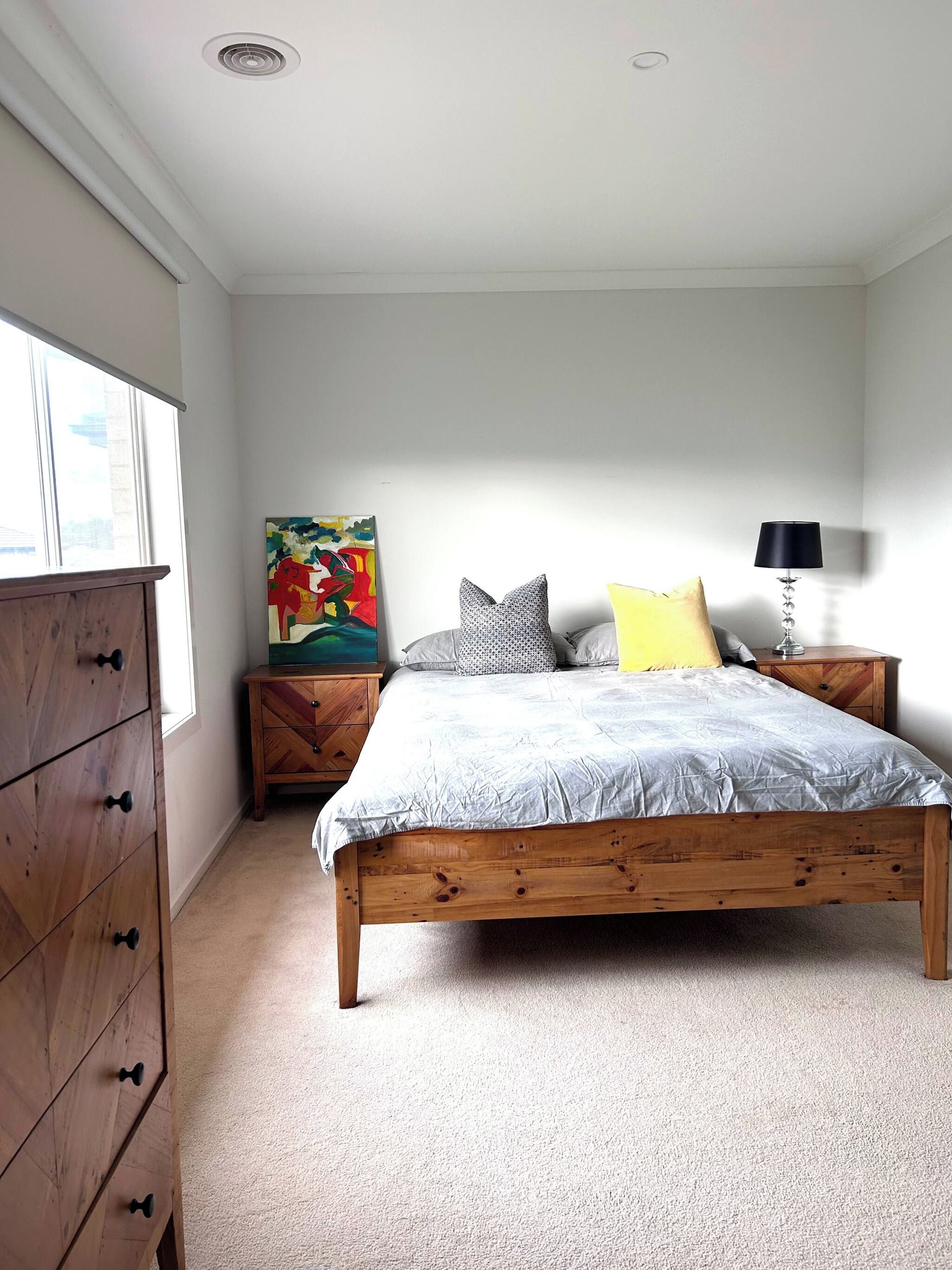 A bedroom with a wooden bed, a nightstand with a lamp, a dresser, and a colorful painting on the wall.