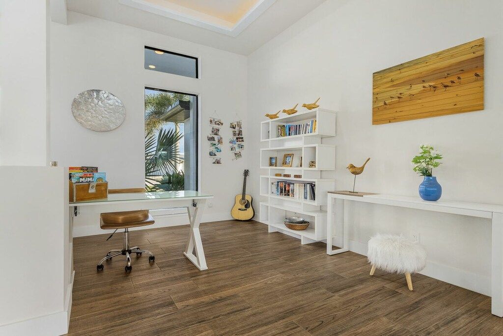 A room with a desk, bookshelves, a guitar, and a window with a view of palm trees.