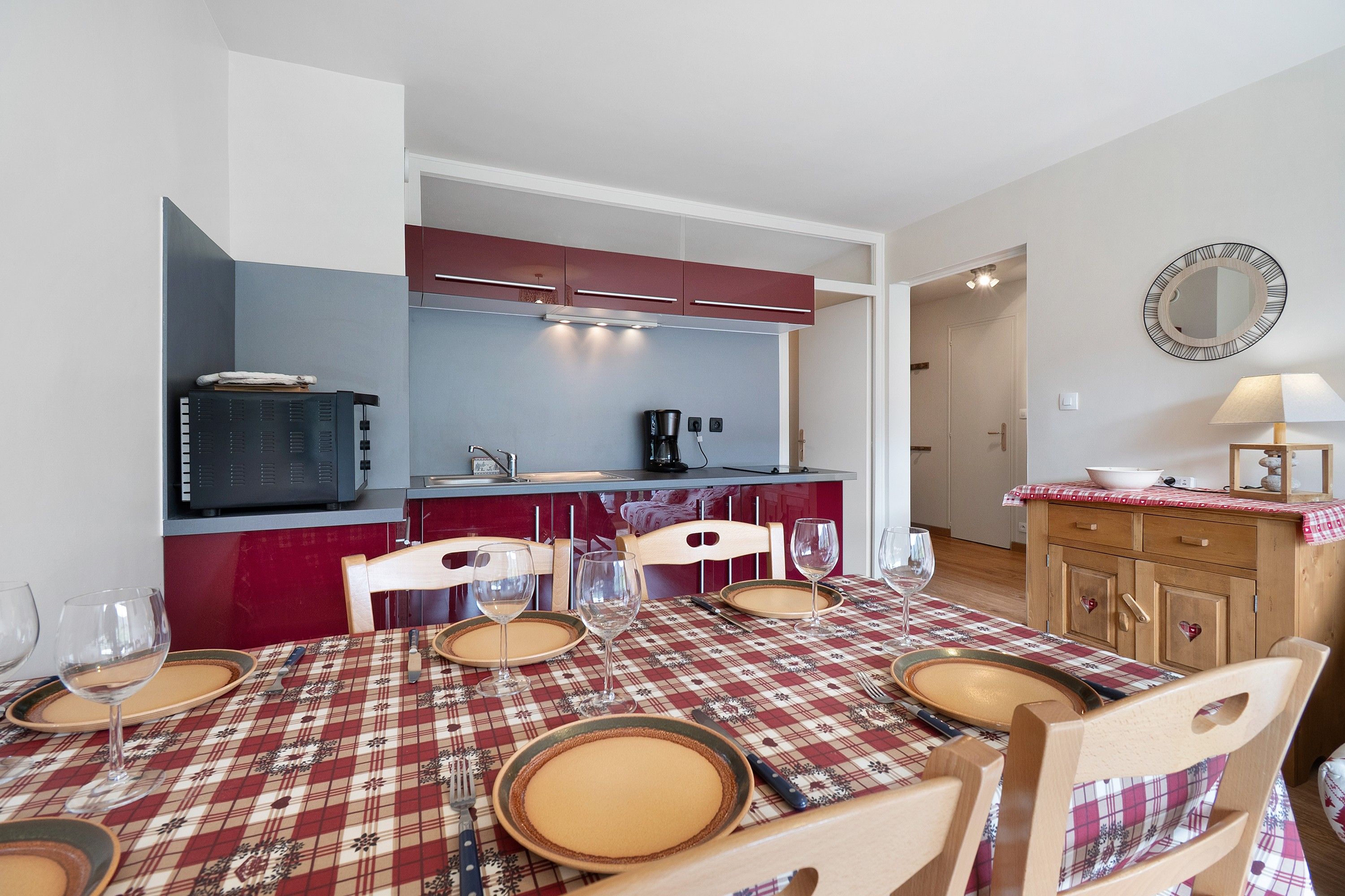 A dining area with a red and white checkered tablecloth, wooden chairs, and a kitchen in the background.