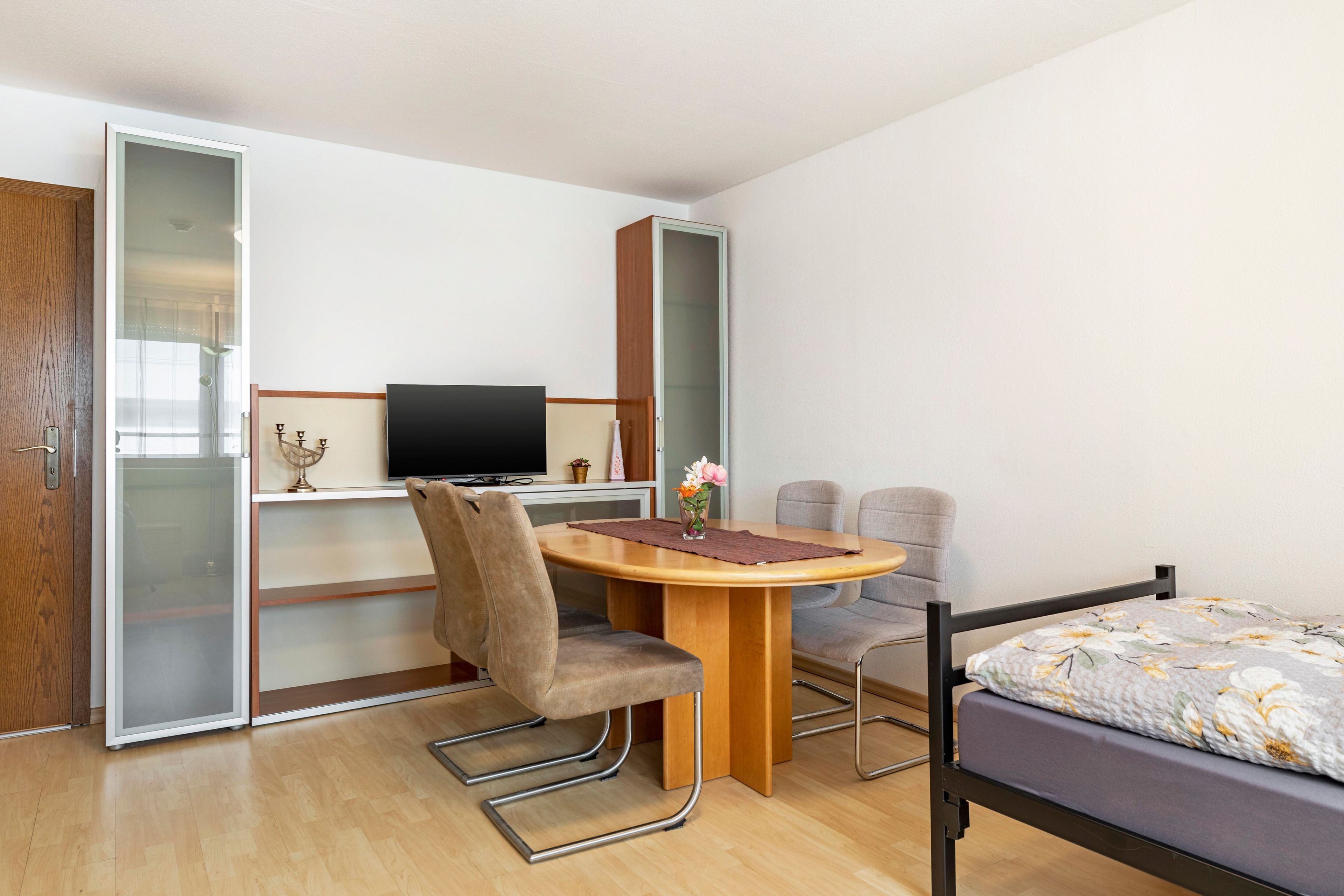 Habitación compacta con cama, escritorio con computadora, silla y una pequeña mesa con un jarrón de flores.