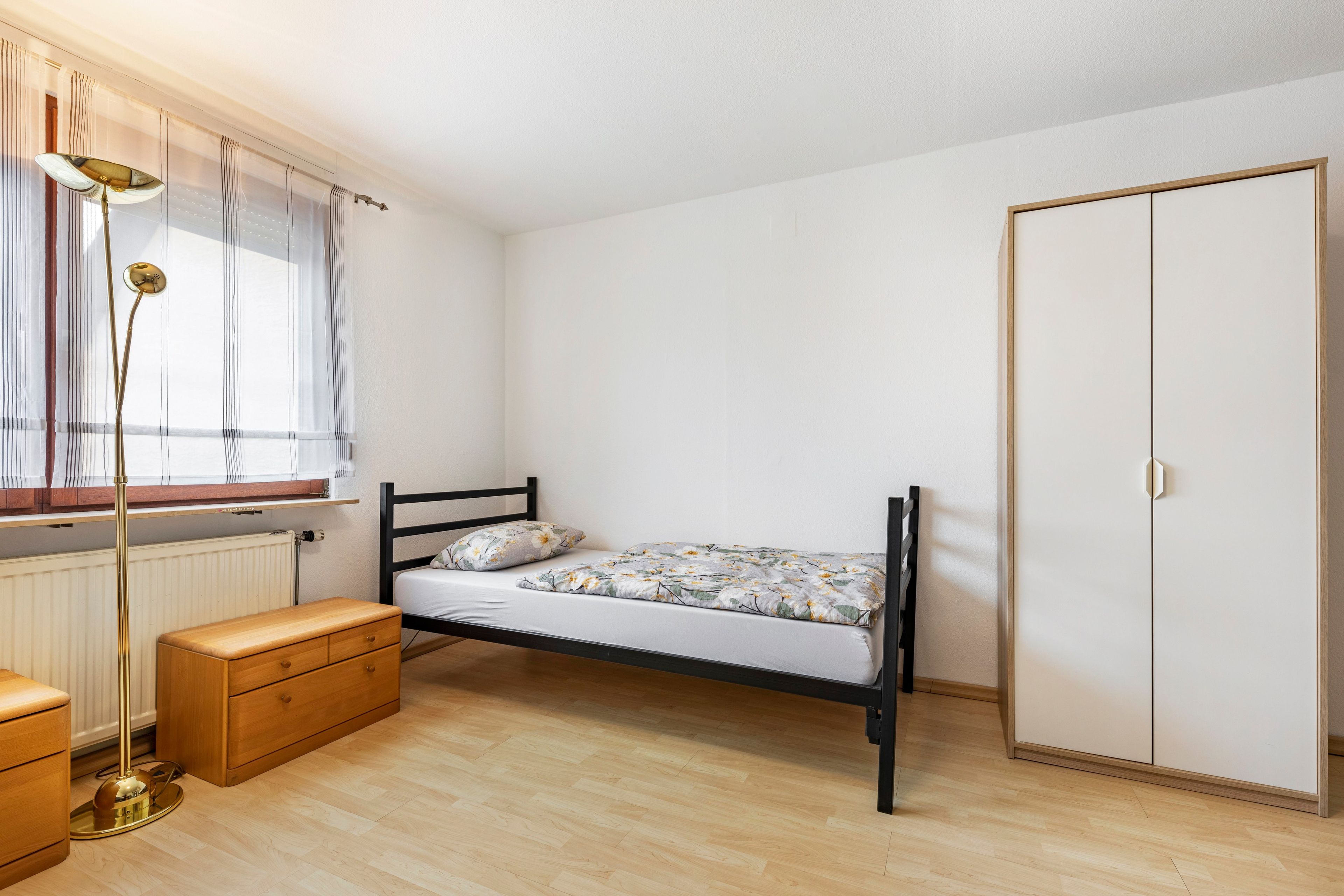 A bedroom with a single bed, a wardrobe, a bedside table, a lamp, and a window with curtains.