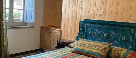 A bedroom with a wooden wall, a bed with a patterned bedspread, a chandelier, and a window with curtains.