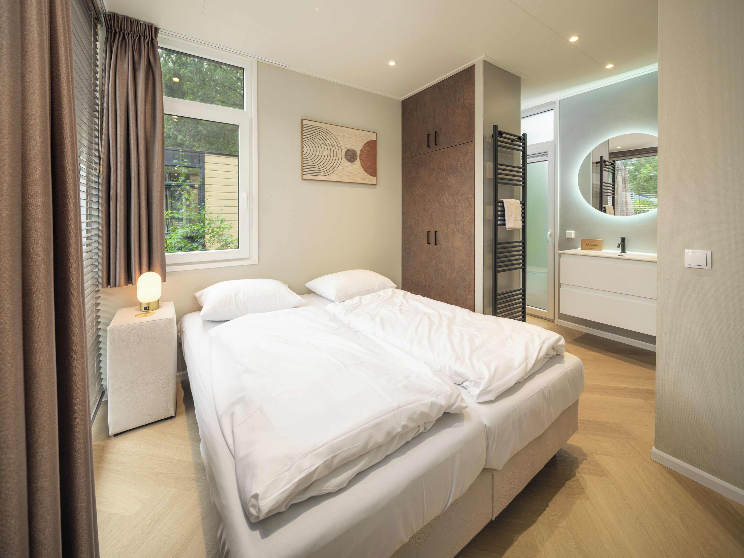 Een moderne slaapkamer met een groot bed, een nachtkastje met een lamp, een kledingkast en een badkamer met een spiegel en wastafel.