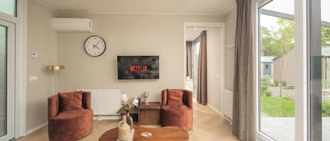 Una sala de estar moderna con un ventanal grande, dos sofás, una mesa de centro, un reloj y un televisor que muestra 'NETFLIX'.