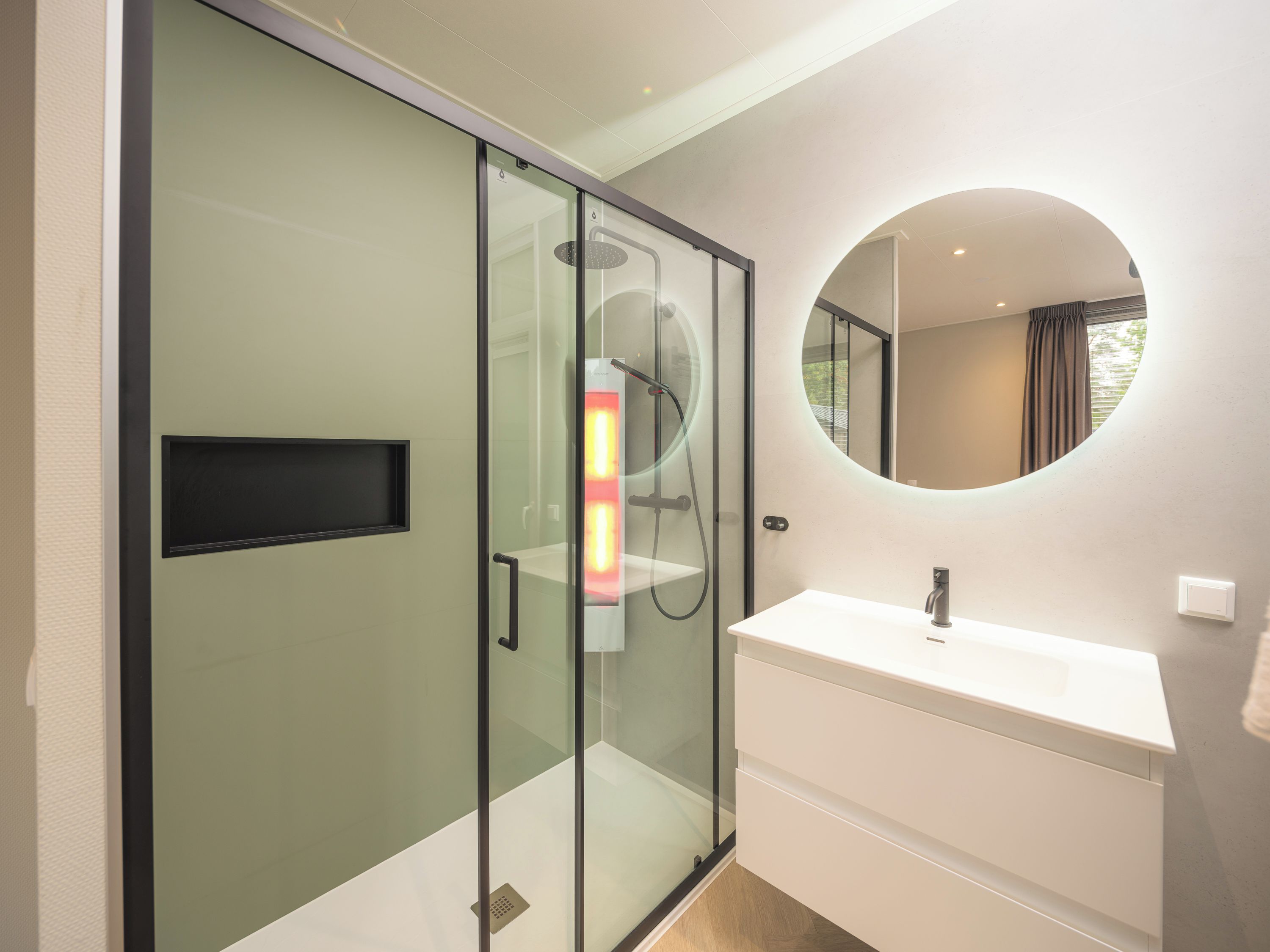 Een moderne badkamer met een douchecabine van glas, een wit wastafelmeubel en een ronde spiegel.