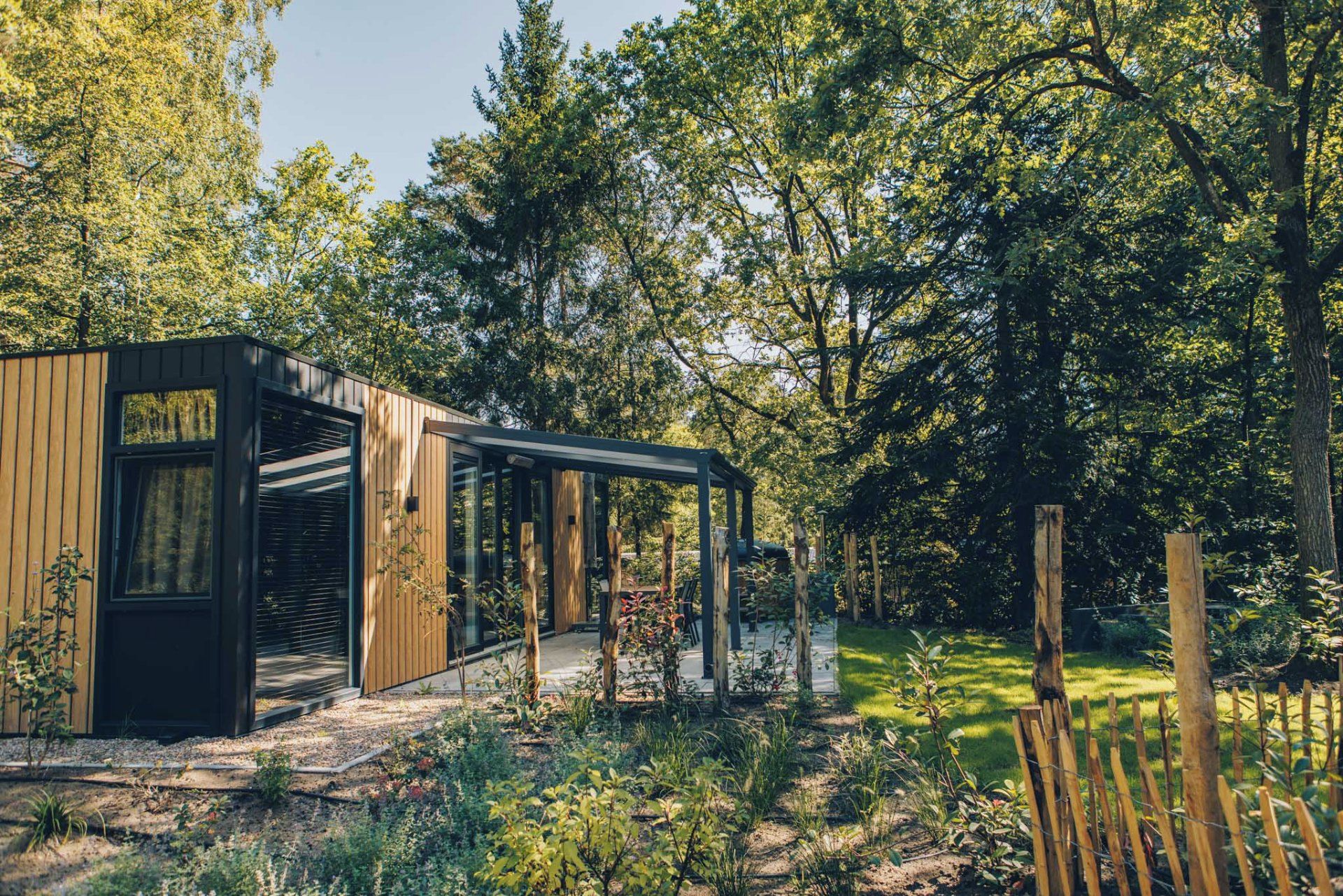 A modern wooden cabin with large windows, surrounded by a lush garden and dense forest.
