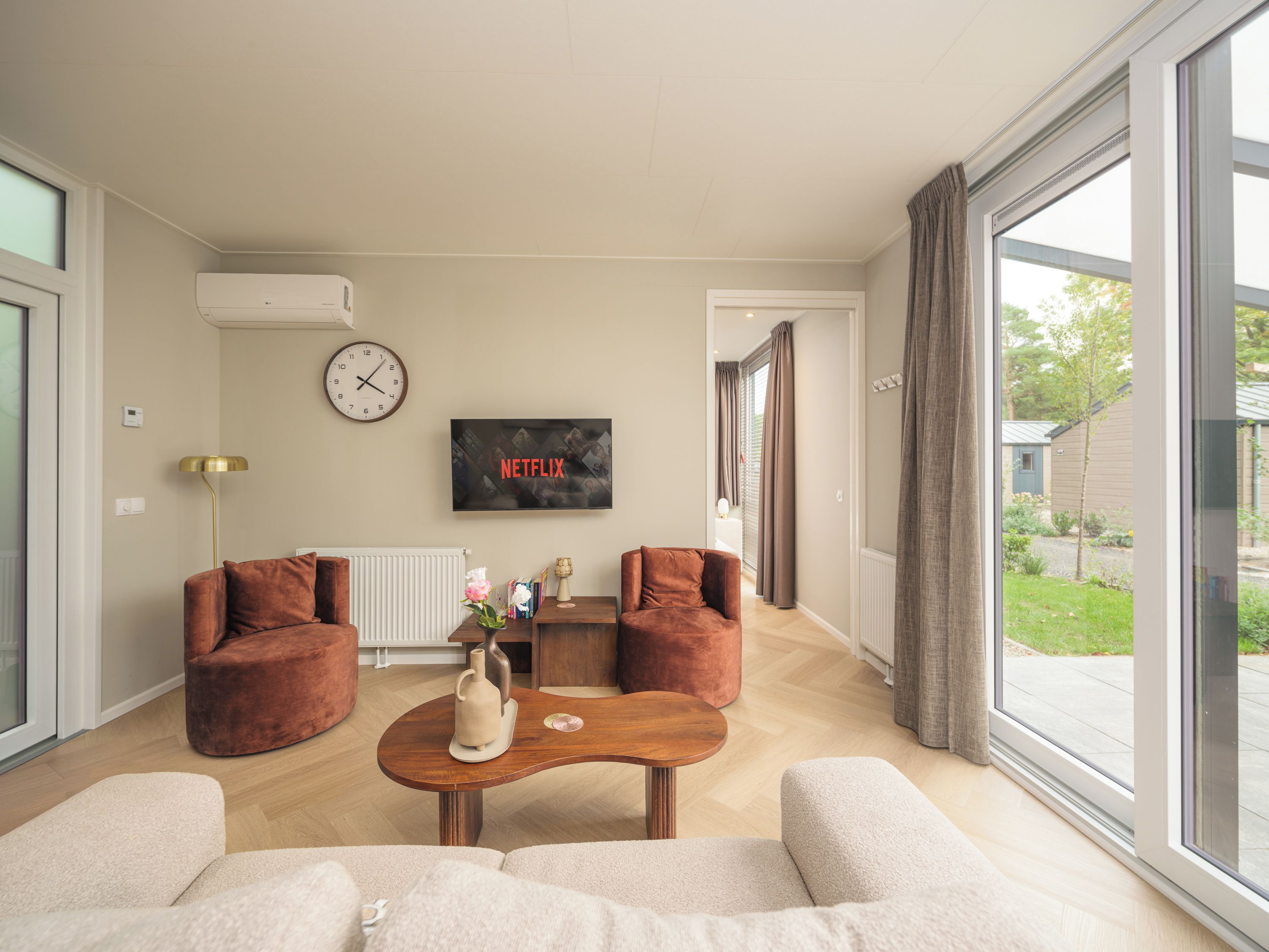 Una sala de estar moderna con un ventanal grande, dos sofás, una mesa de centro, un reloj y un televisor que muestra 'NETFLIX'.