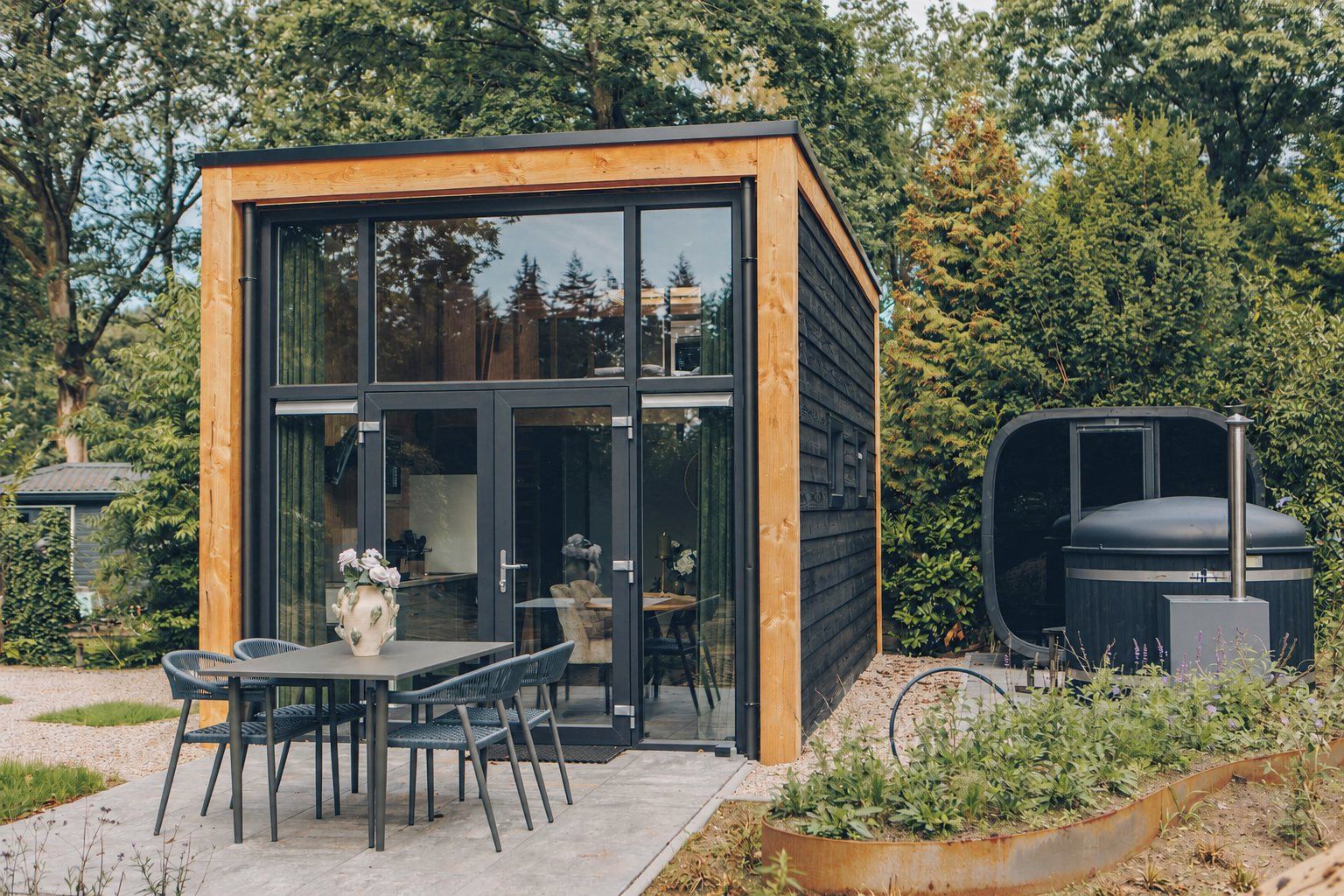 Uma cabana de madeira moderna com porta de vidro, um alpendre com mesa e cadeiras, e uma churrasqueira.