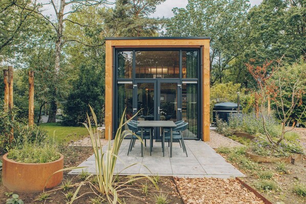 Ein moderner Essbereich im Freien mit Glasveranda, umgeben von einem Garten mit verschiedenen Pflanzen und Bäumen.