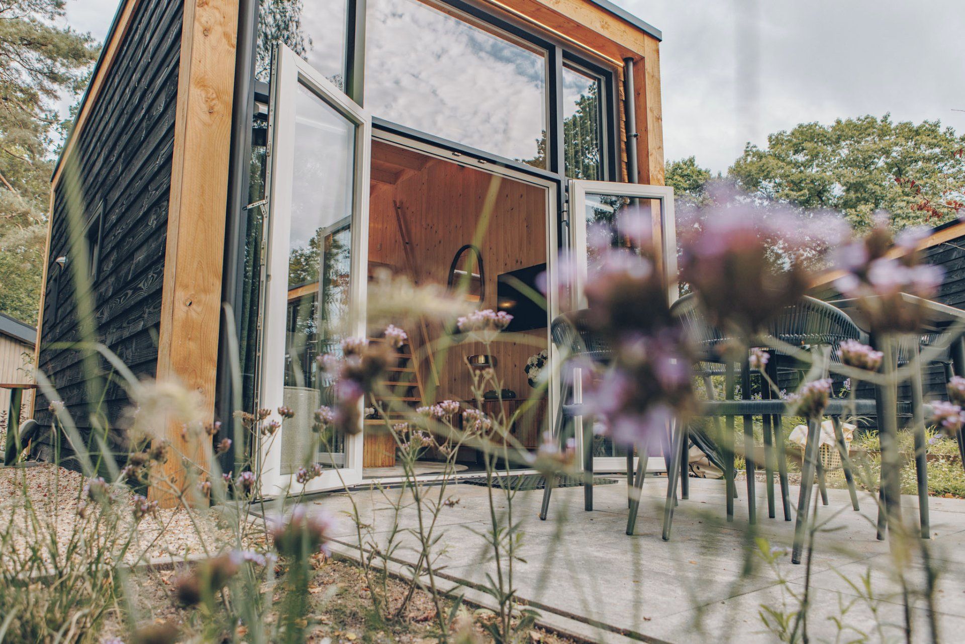 Une cabane moderne en bois, dotée d’une porte en verre, d’une terrasse en béton et d’un jardin planté de fleurs sauvages.