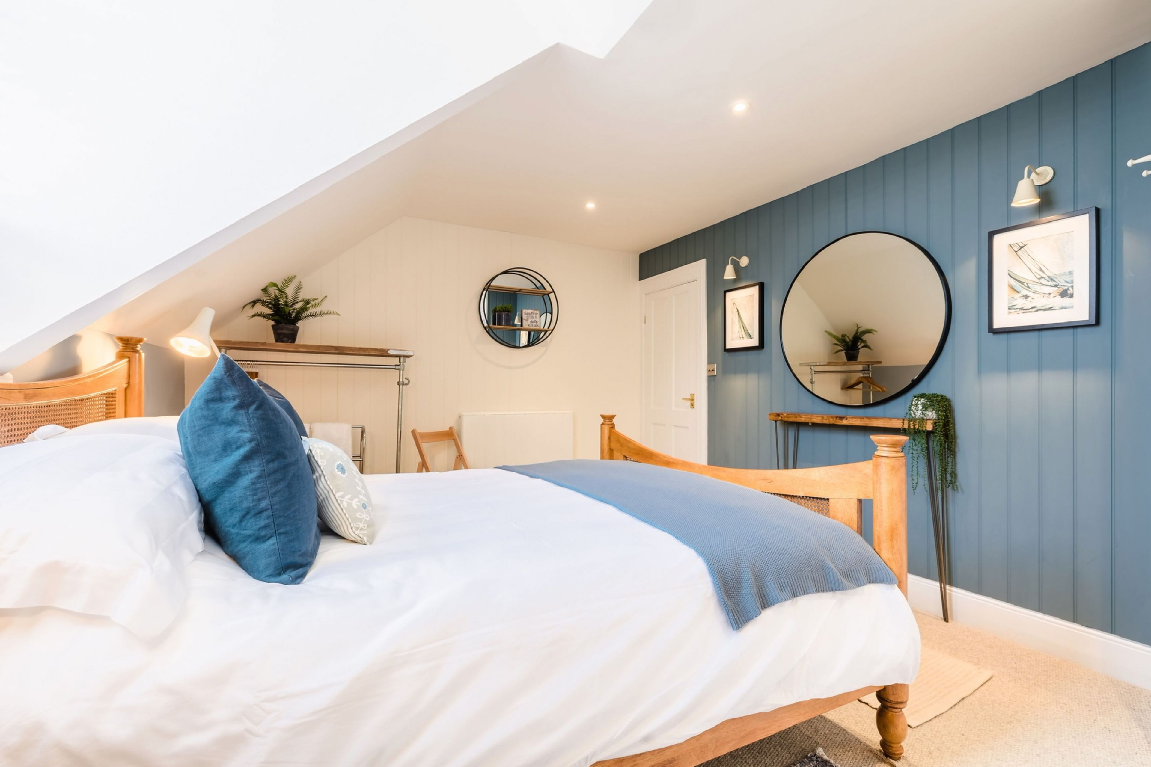 Un dormitorio con techo inclinado, cama con ropa de cama blanca y azul, cabecero de madera, espejo redondo y una pared de acento azul.