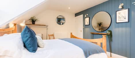 Un dormitorio con techo inclinado, cama con ropa de cama blanca y azul, cabecero de madera, espejo redondo y una pared de acento azul.