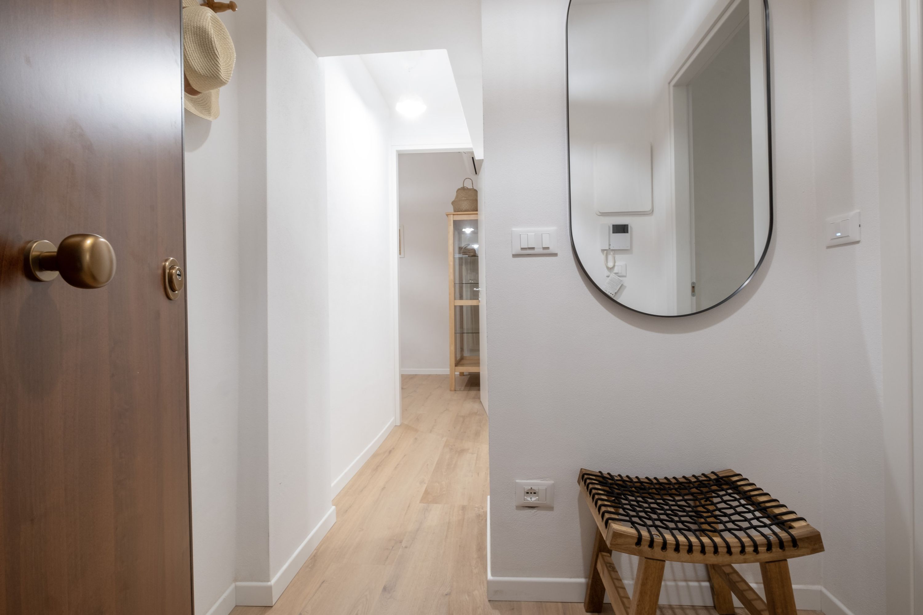 Un pasillo con una puerta de madera, un espejo redondo y un taburete de madera.