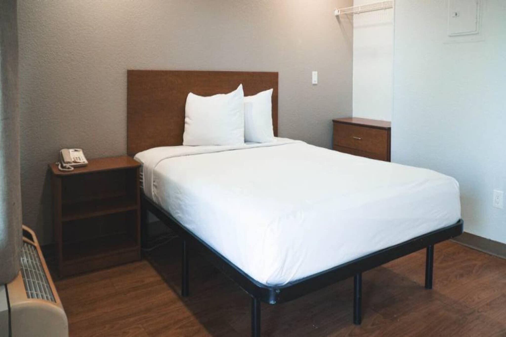 Quarto de hotel com uma cama de solteiro, mesa de cabeceira com telefone e um criado-mudo de madeira.