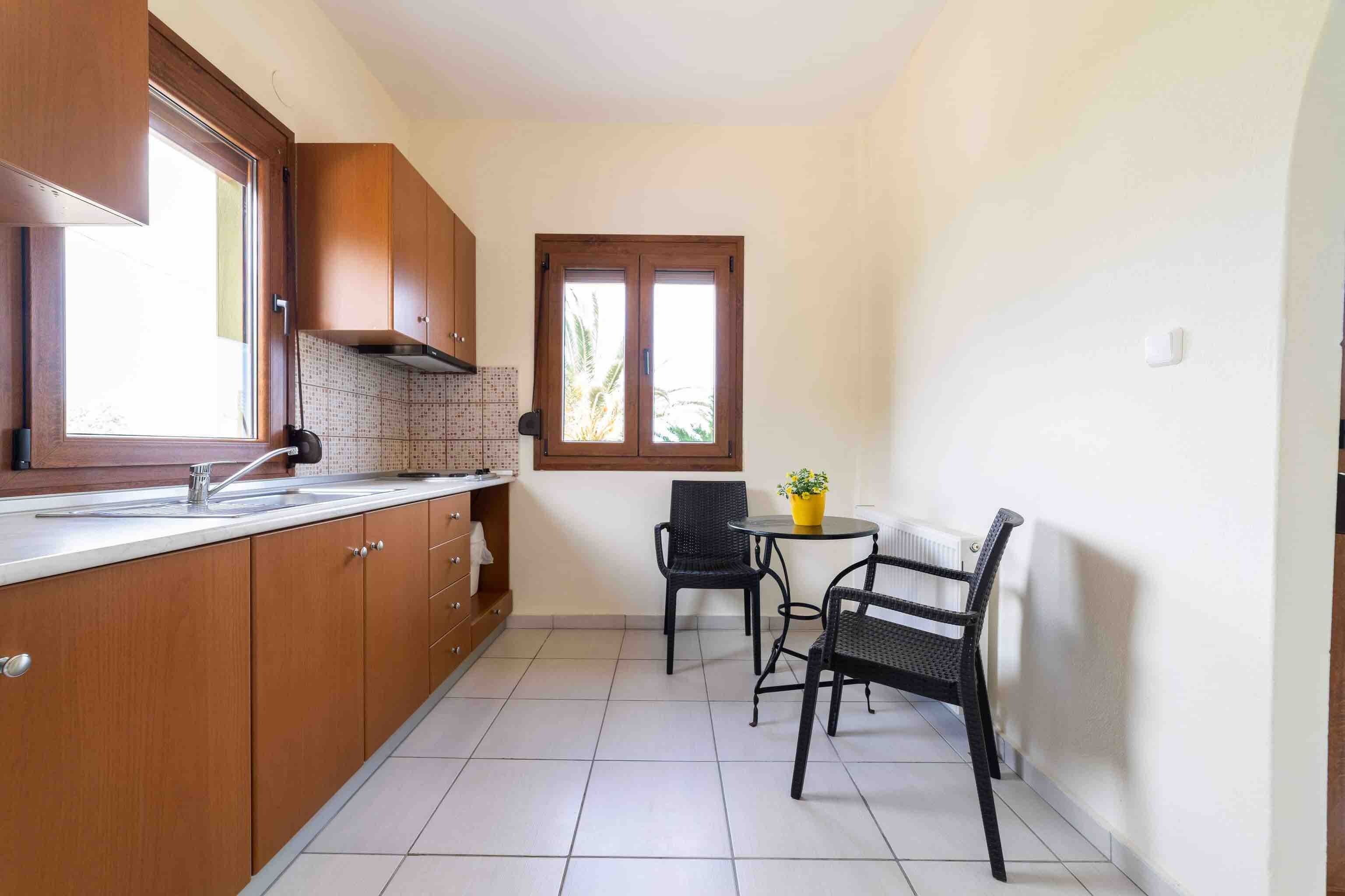 Een knusse keuken met houten kastjes, een betegelde achterwand, een raam en een kleine eethoek met twee stoelen en een tafel.
