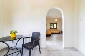 Une pièce comprenant une table à manger et des chaises, un radiateur, et une arche donnant sur un espace de vie avec un canapé et une fenêtre.