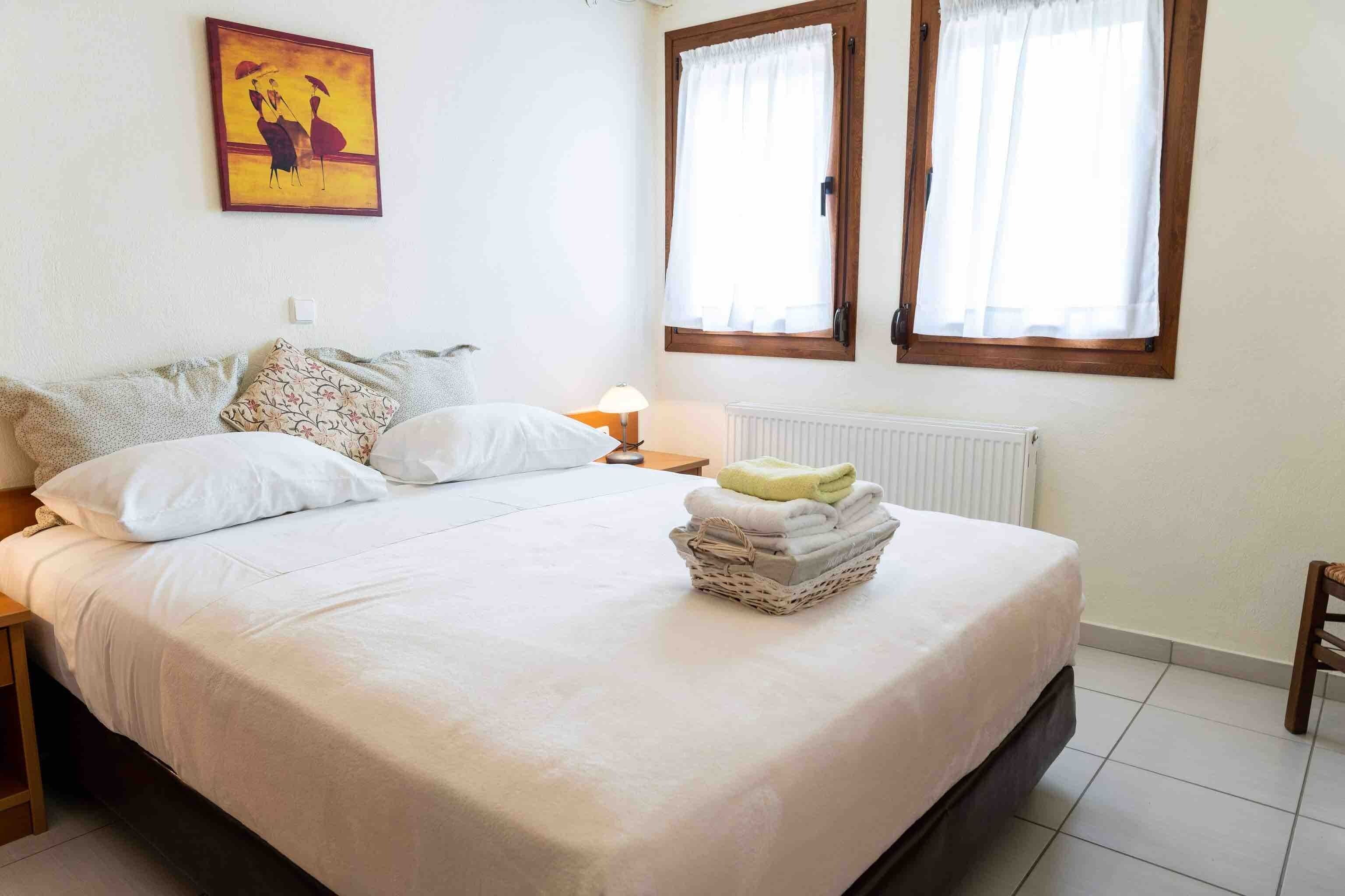 Een slaapkamer met een bed, kussens, een nachtkastje met een lamp, een radiator en een raam met gordijnen.