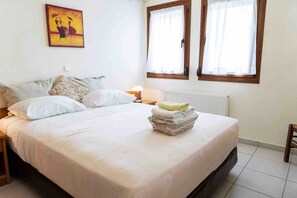Une chambre à coucher avec un lit, des oreillers, une table de chevet avec une lampe, un radiateur et une fenêtre avec des rideaux.