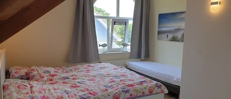 A bedroom with a bed, a window with curtains, a wall-mounted light, and a beach-themed painting.