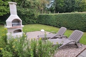 An outdoor seating area with a stone-built barbecue grill, surrounded by a hedge and gravel. - Holiday paradise sea air (Kamperland)