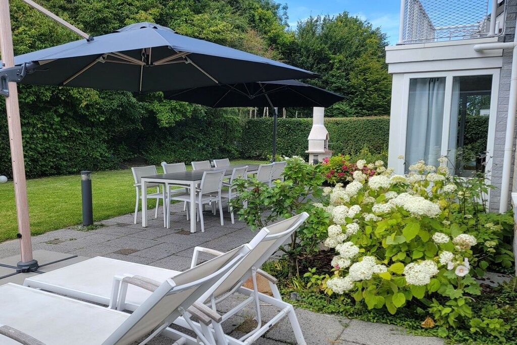 Een terras buiten met witte stoelen en een tafel, een grote parasol, en een tuin met witte bloemen.