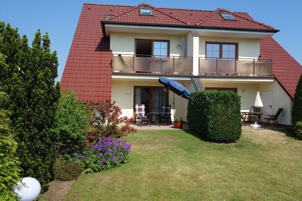 Ein zweistöckiges Wohnhaus mit rotem Ziegeldach, einem Balkon im oberen Geschoss und einem kleinen Garten mit Rasenfläche und Topfpflanzen.