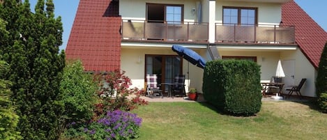 Ein zweistöckiges Wohnhaus mit rotem Ziegeldach, einem Balkon im oberen Geschoss und einem kleinen Garten mit Rasenfläche und Topfpflanzen.