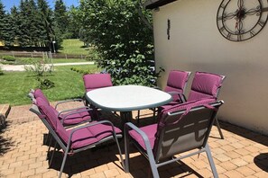 Eine Terrasse im Freien mit einem runden Tisch und vier Stühlen, umgeben von einem Pflasterboden und einem Garten mit Bäumen und einem Zaun.