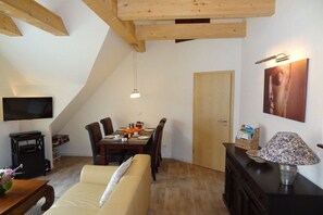 Ein gemütliches Wohnzimmer mit einer Couch, einem Esstisch und Stühlen. Ein Fernseher hängt an der Wand, eine Lampe steht auf einem Beistelltisch und es gibt eine Holzzieldecke.